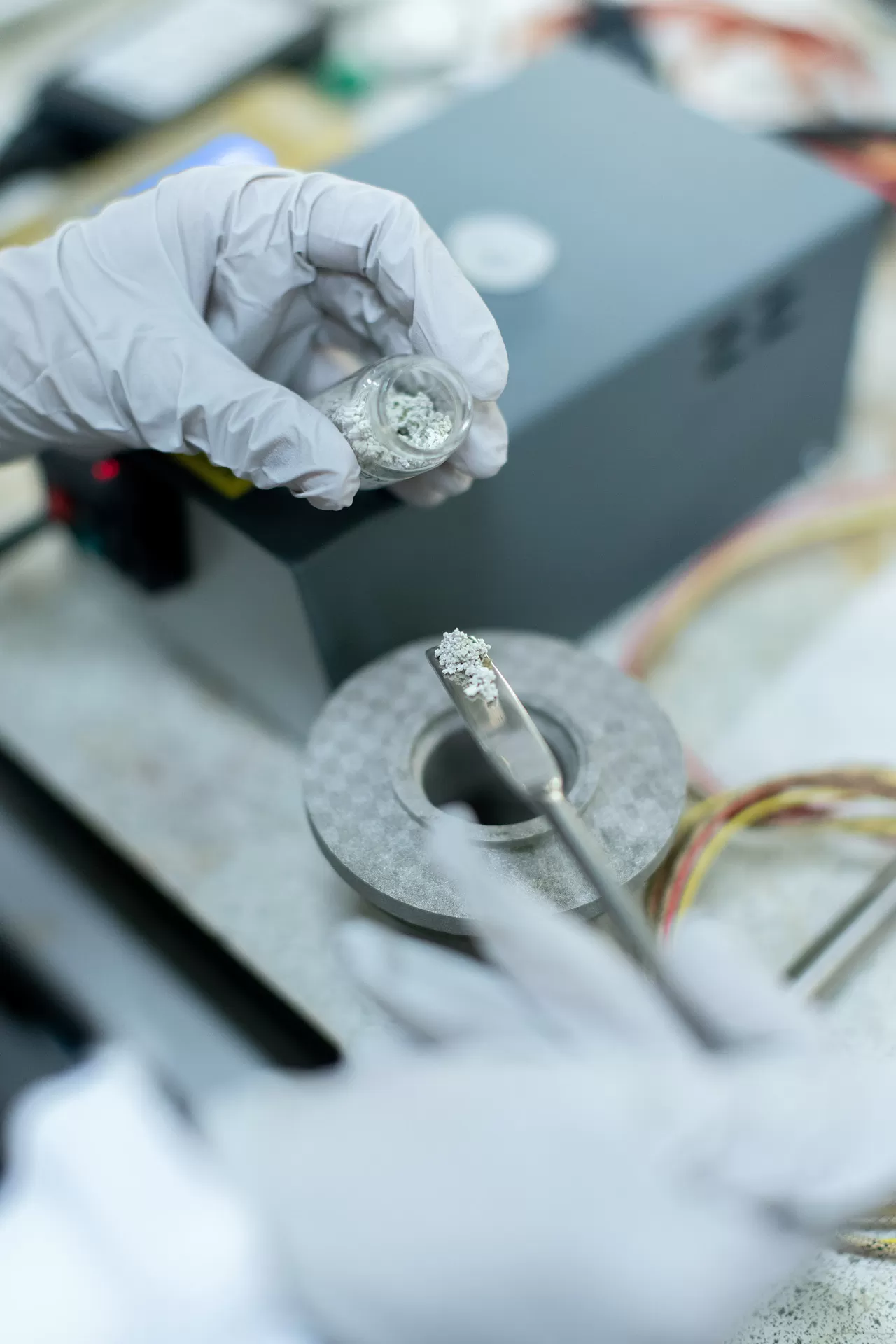Nahaufnahme von Händen mit Laborhandschuhen, die Pulver auf Metalllöffel abmisst. Eine Ingenieurin entwickelt saubere Energiespeicherlösungen.