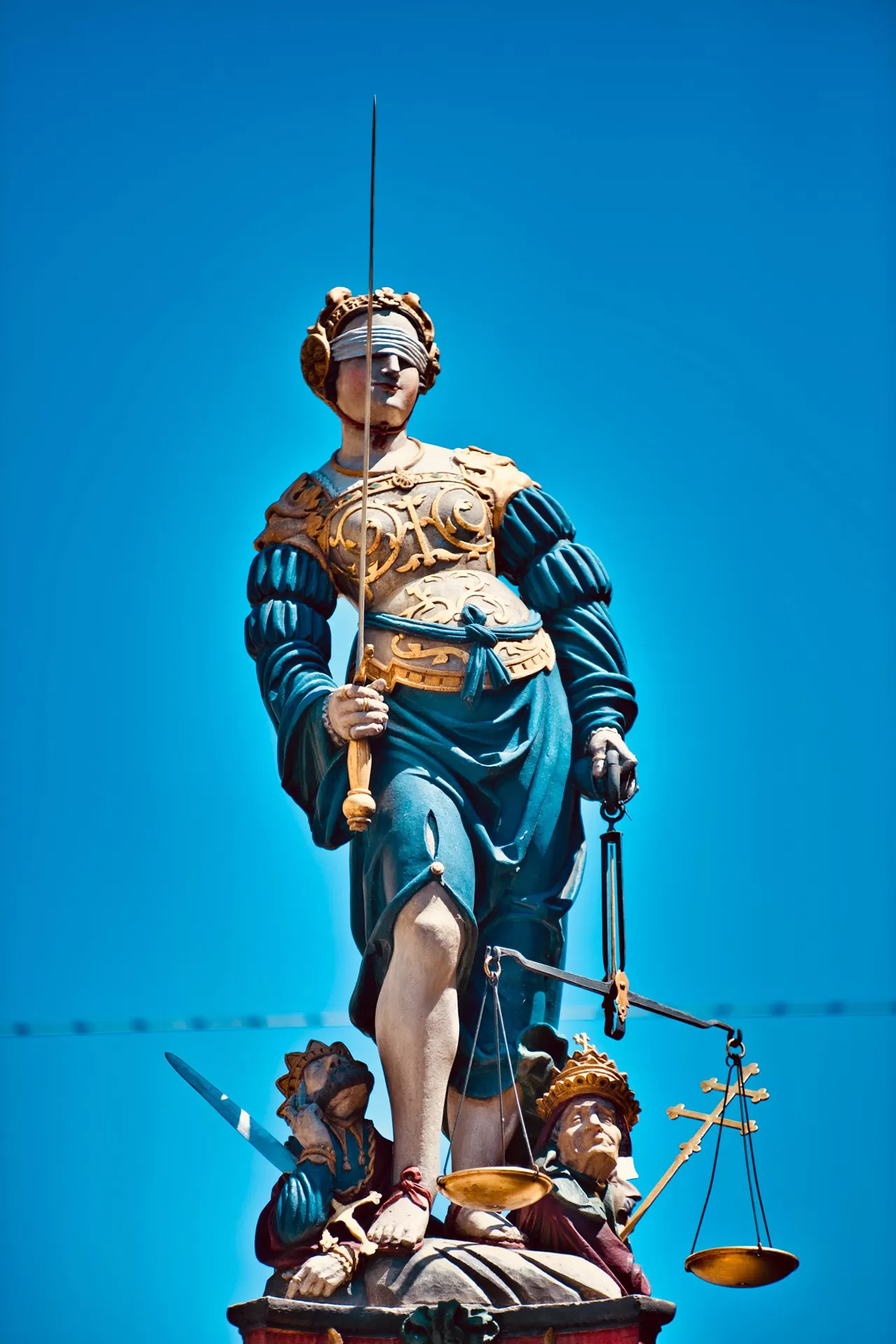 Eine Statue der Justizia in blauem Gewand und mit goldener Rüstung steht auf dem Gerechtigkeitsbrunnen in Bern. In den Händen hält sie Schwert und Waage, die Augen sind verbunden.