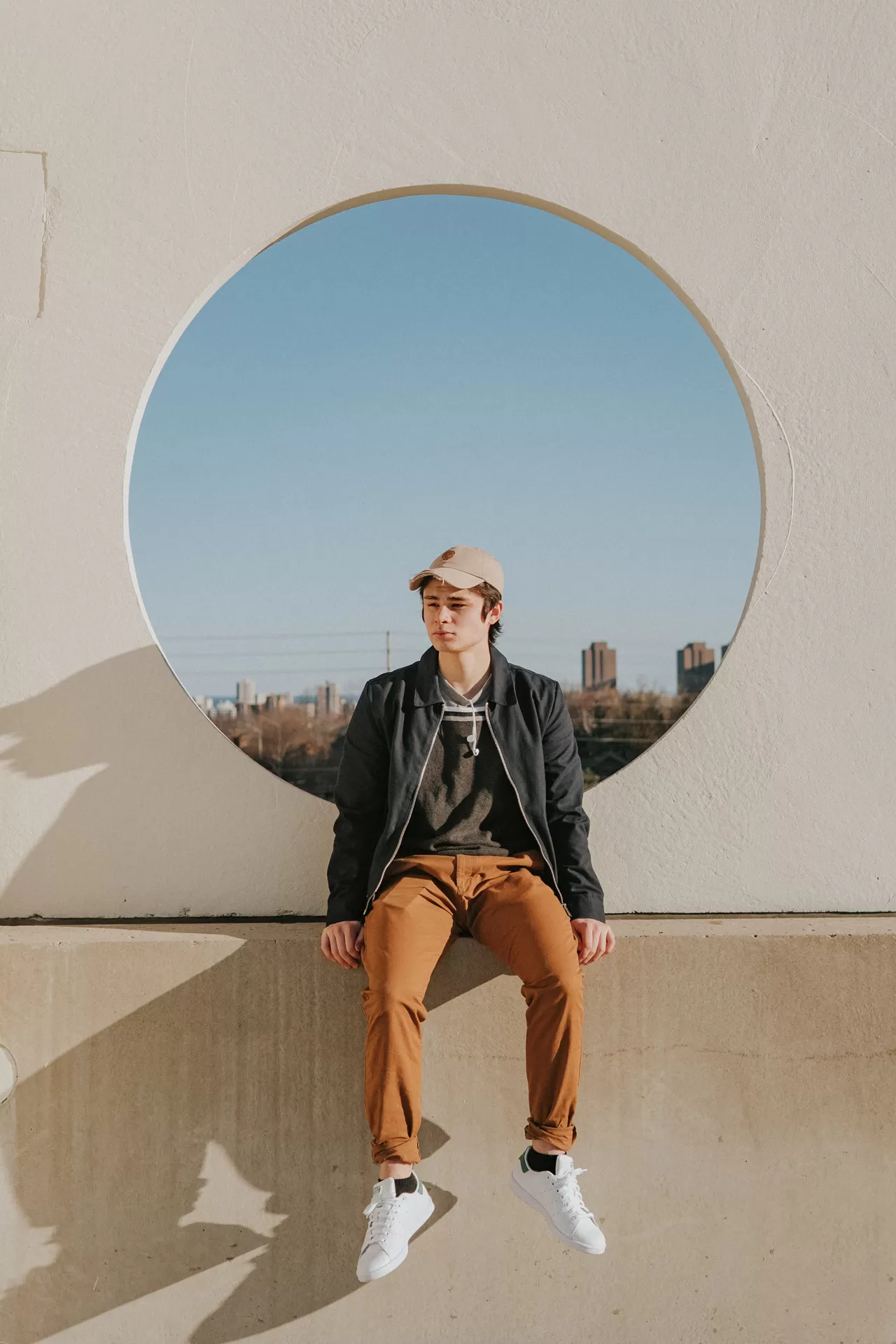 Nachdenklicher Teenager sitzt auf Betonmauer, hinter ihm ein kreisrundes Fensterloch mit Blick auf eine Stadt.