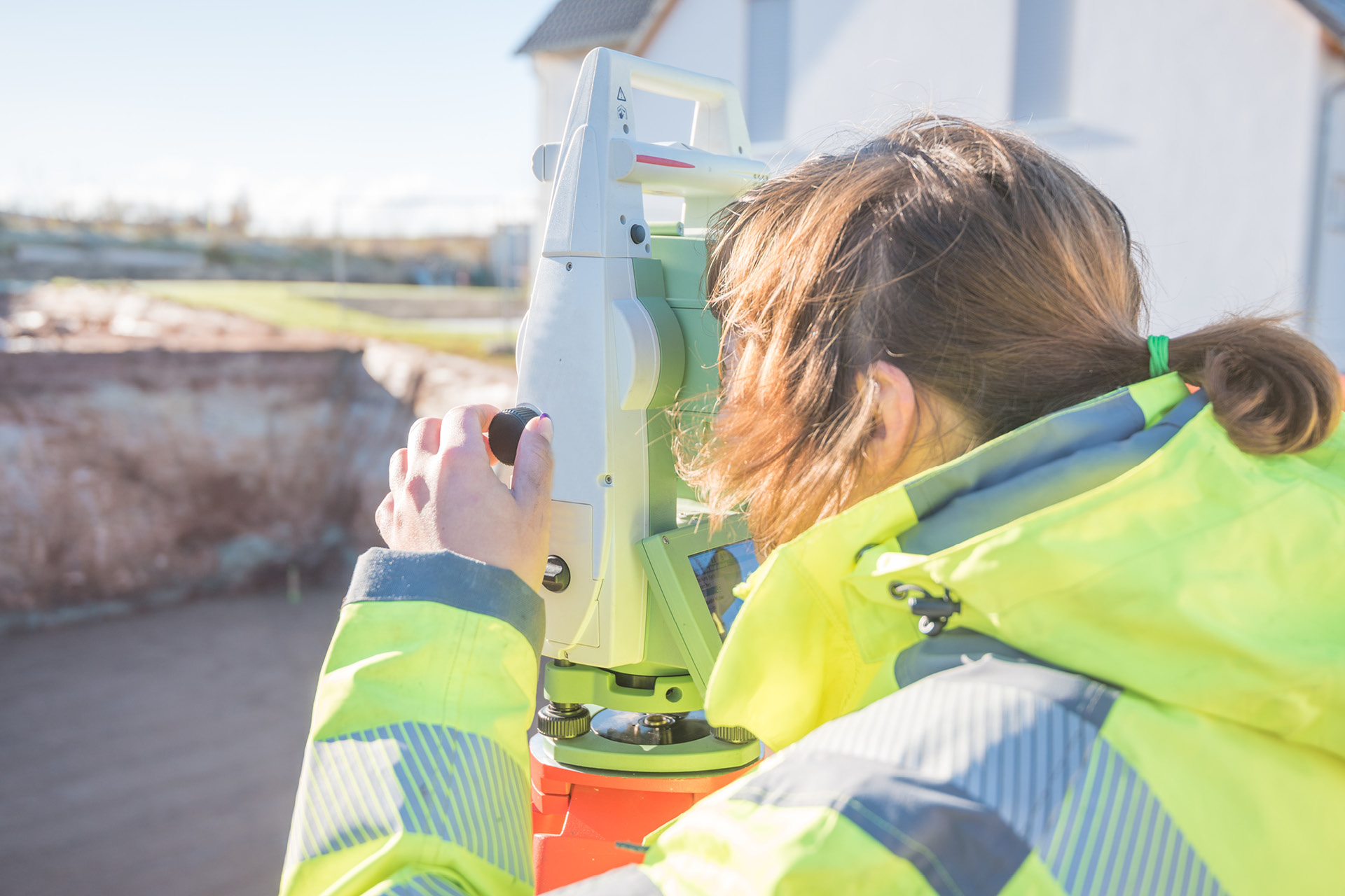 Frau in gelber Schutzjacke bedient Tachymeter zur Gebäudevermessung beim Hausbau.