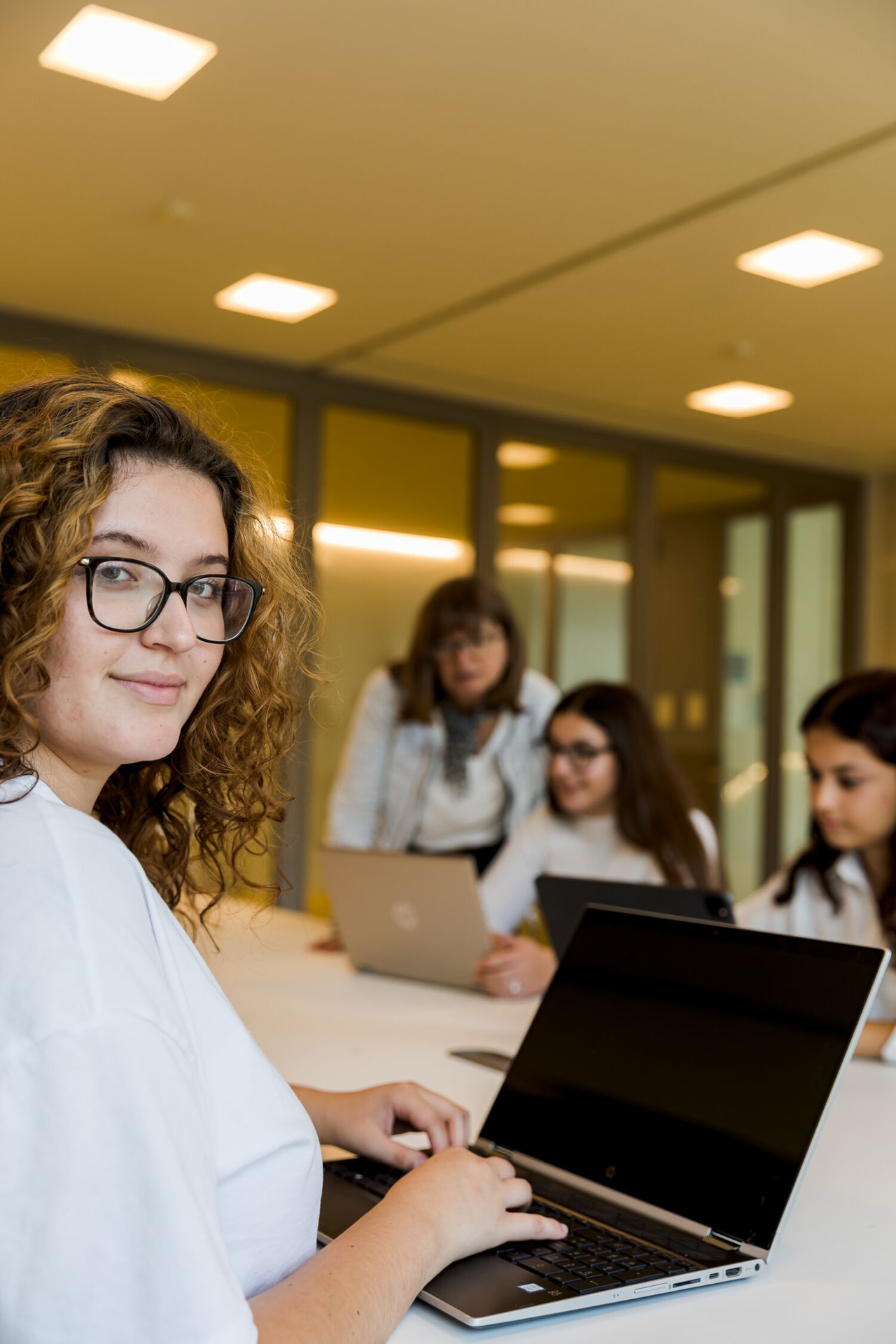 Jugendliche Schülerinnen arbeiten am Laptop und werden von der Lehrerin unterstützt. Eine Schülerin schaut direkt zum Betrachter.