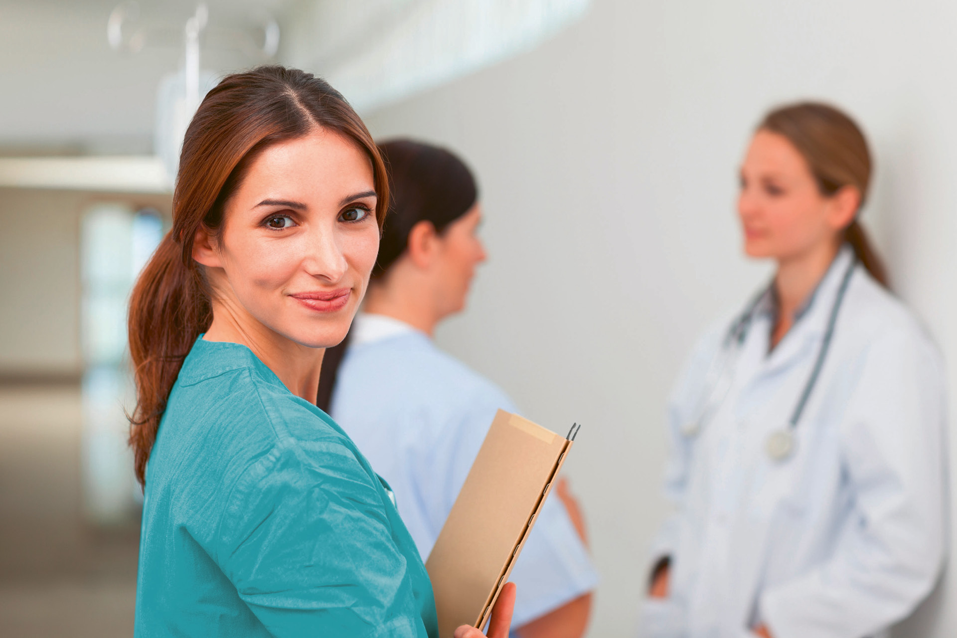 Drei Gesundheitsfachpersonen stehen im Flur eines Spitals. Zwei unterhalten sich, die dritte Person hat ein Dossier in der Hand und schaut in die Kamera.