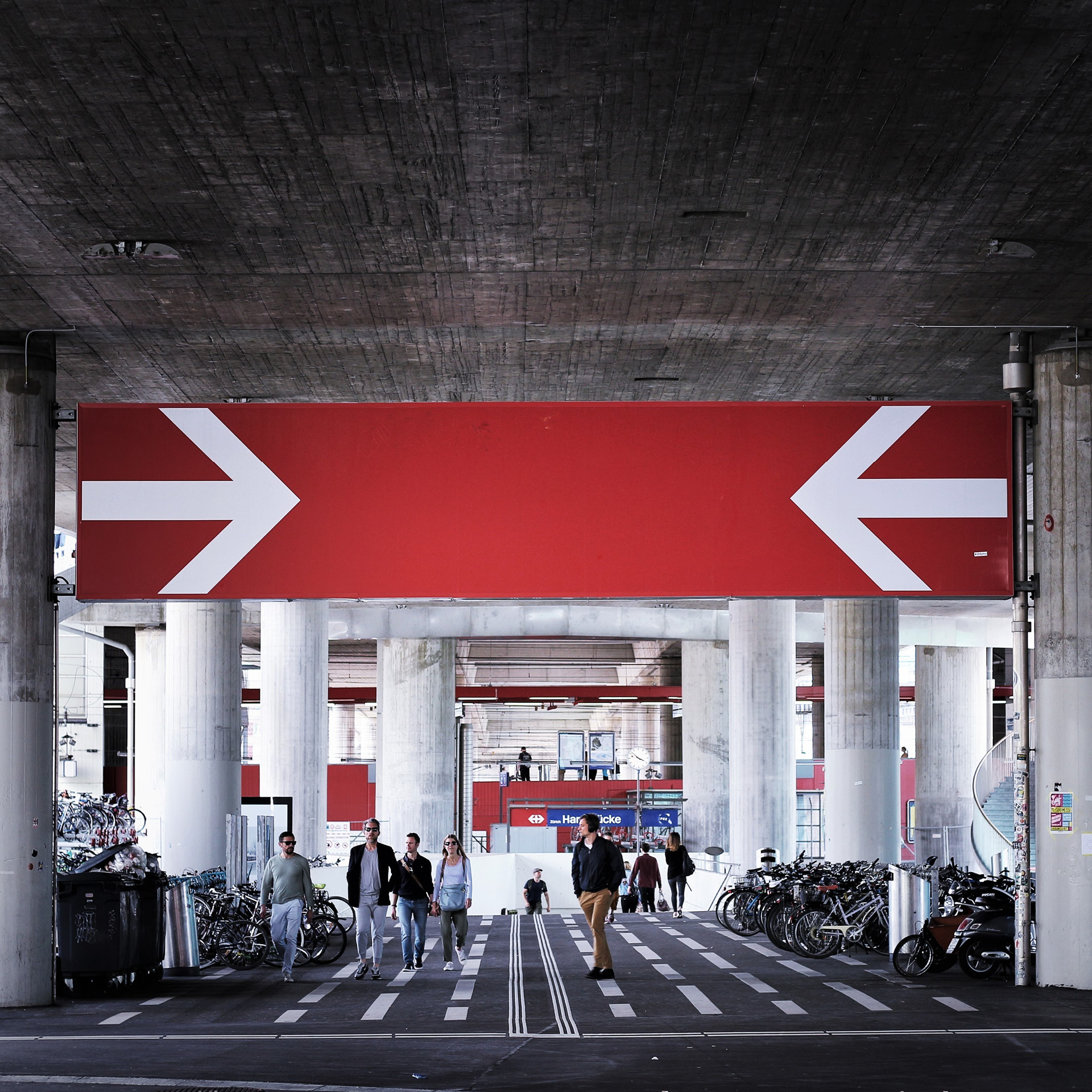 Am Bahnhof Hardbrücke in Zürich kommen Leute aus der Unterführung. Im Hintergrund erkennt man die Perrons der Züge, über die Zugstrecke kreuzt eine grosse Strassenbrücke.
